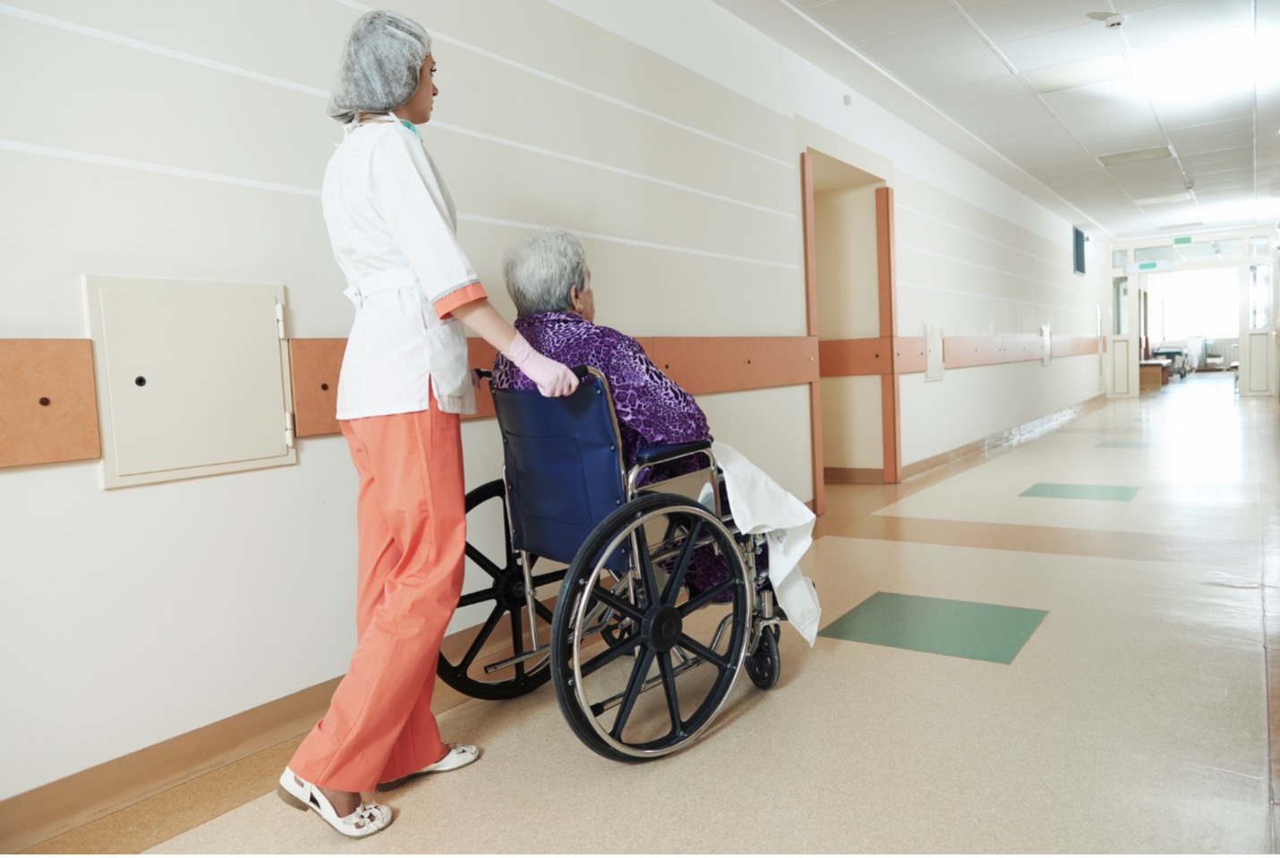 Nurse pushing elderly patient in wheelchair down facility hallway